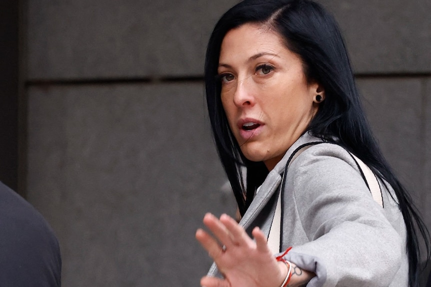 A woman wearing a suit and walking outdoors looks at the camera and raises her hand