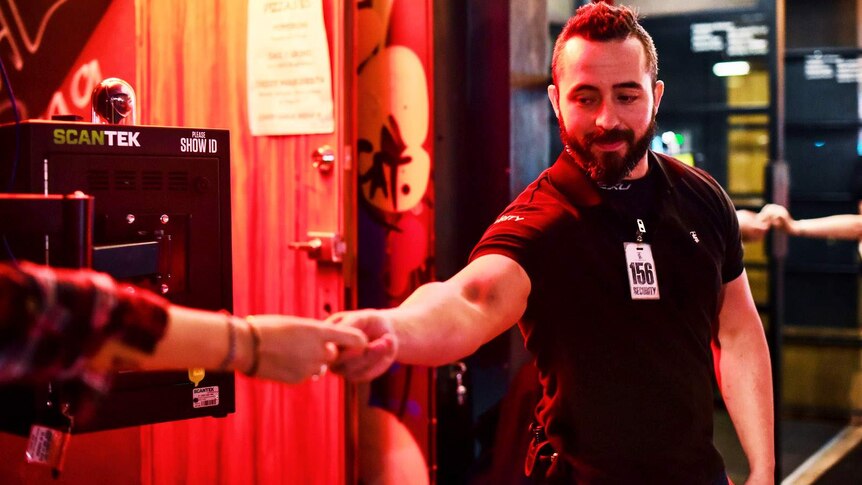 A bouncer standing next to an ID scanner takes ID off patron.