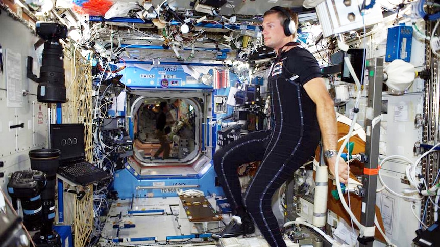 a man in a garment akin to a wetsuit leans against a wall of machinery inside a space station.