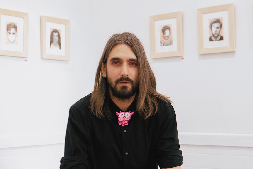 A white person with long hair and a beard sits in a chair in an art gallery and leans forward, clasping their hands