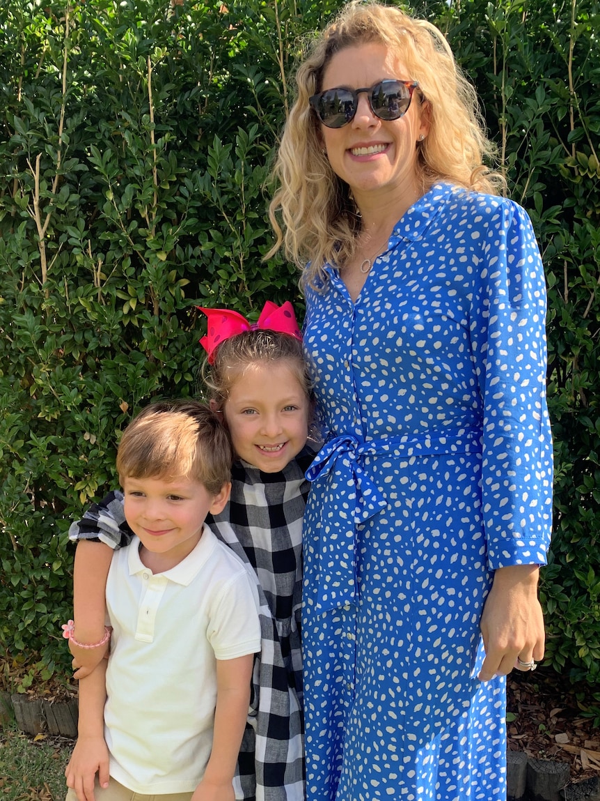 Two children and a woman stand in front of a hedge.