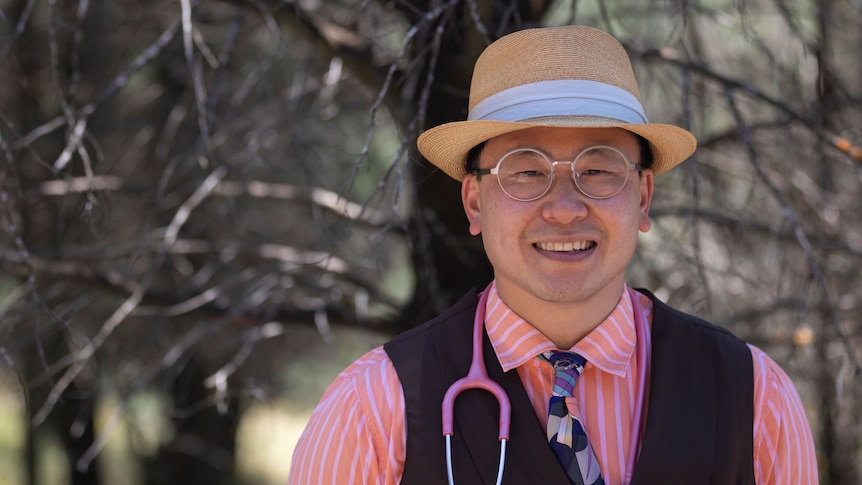 A man in a tan hat, glasses, pale pink shirt and waistcoat stands in front of trees and smiles with a stethoscope on his neck.