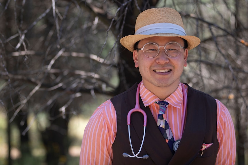 A man in a tan hat, glasses, pale pink shirt and waistcoat stands in front of trees and smiles with a stethoscope on his neck.