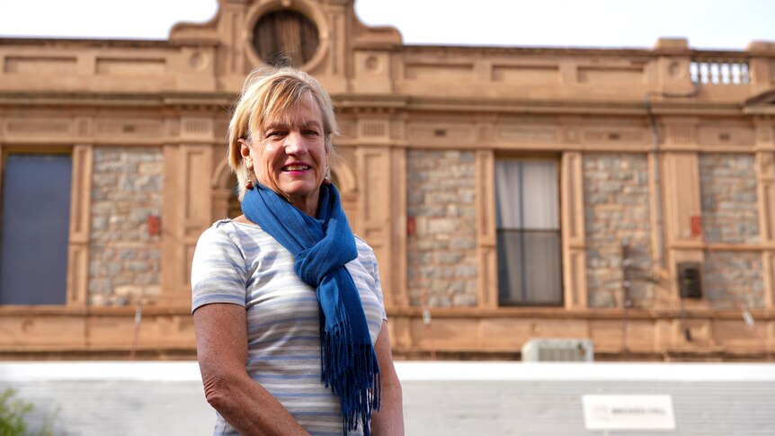 A white woman with a white shirt and blue scarf standing in front of an old building
