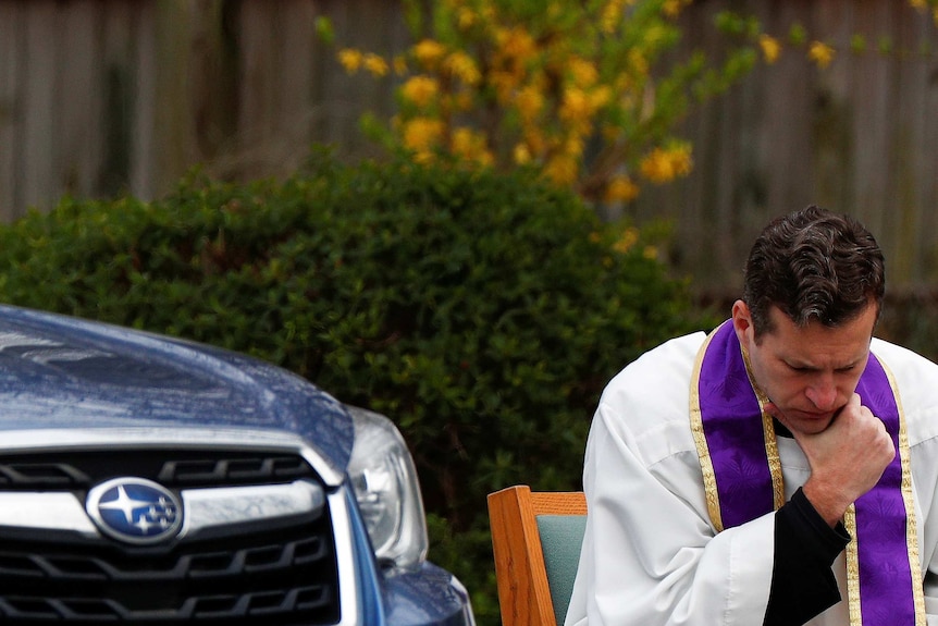 A priest with his head bowed next to a car
