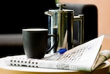 A newspaper open at the crossword sits on a coffee table with a plunger pot and mug, couch in background.