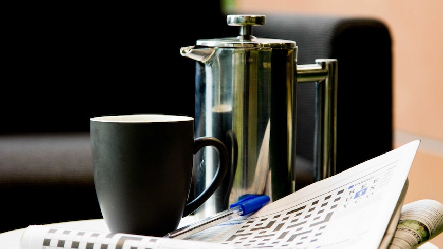 A newspaper open at the crossword sits on a coffee table with a plunger pot and mug, couch in background.