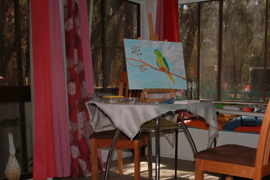 An easel set up on an table. The painting is of a yellow-bellied parrot. Prominent purple curtains. 