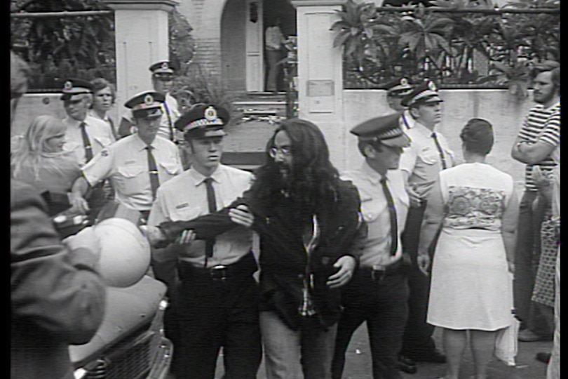 A young man with long hair and a beard being walked out of a house by two police officers.
