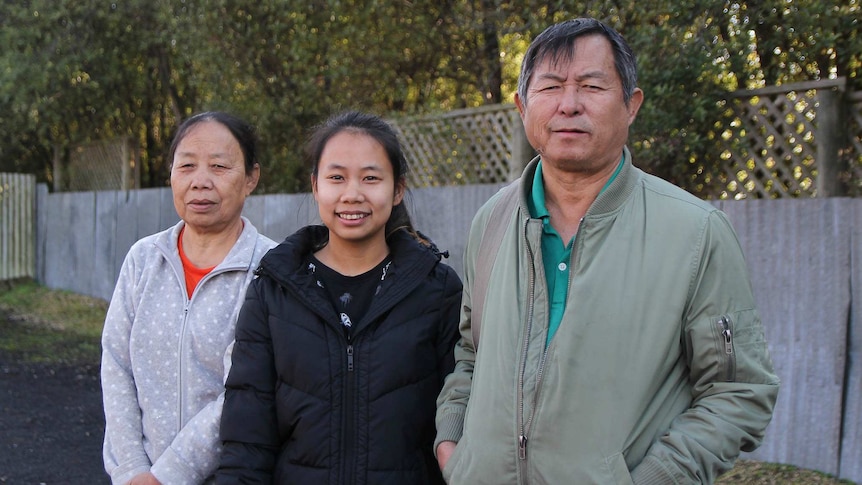 A young woman stands, smiling, between her parents.