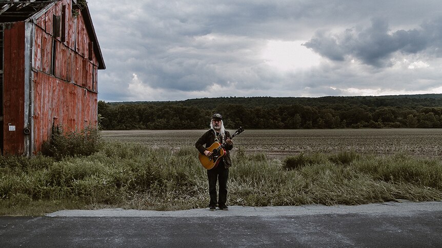 J Mascis stands in front of a barn in an empty field holding an acoustic guitar