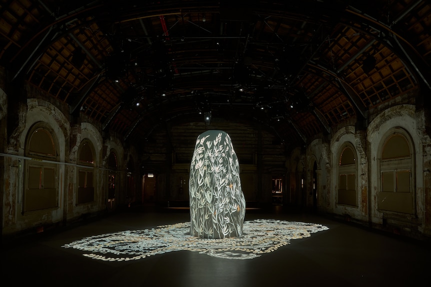 A tall oval shaped object in the middle of a dark warehouse, with illuminated images of flowers and branches projected onto it.