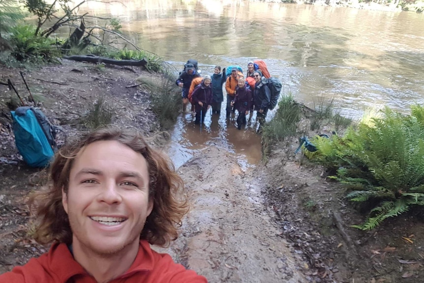 A man close to the camera smiles with a group of seven men and women further back, dressed in trekking gear standing in a river.