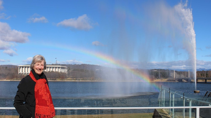 A woman smiles in front of Lake Burley Griffin, with the Captain Cook jet casting a rainbow in the background.