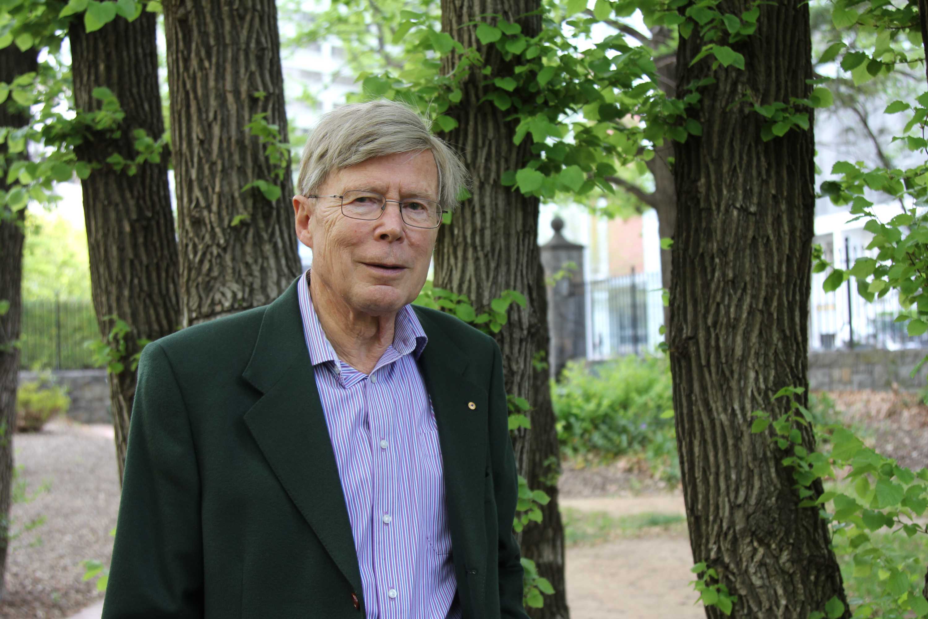 Professor Kurt Lambeck stands in a park, in front of some trees.
