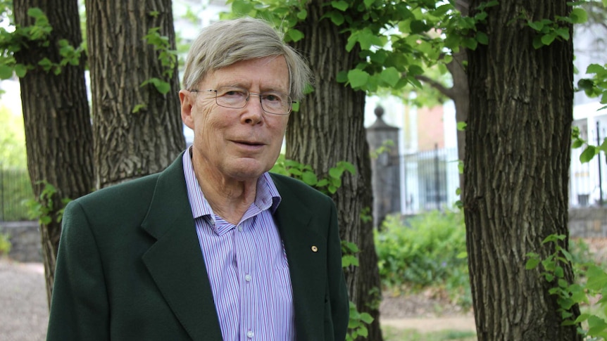 Professor Kurt Lambeck stands in a park, in front of some trees.