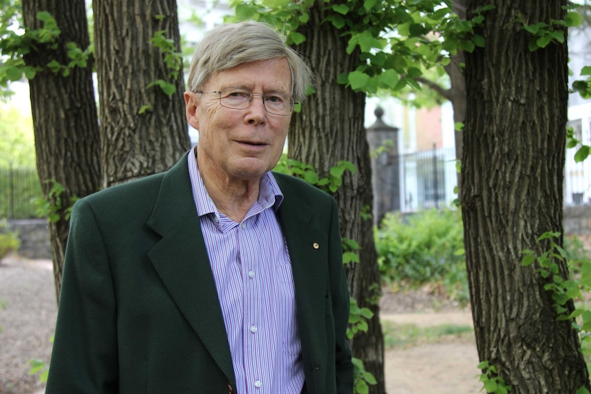 Professor Kurt Lambeck stands in a park, in front of some trees.