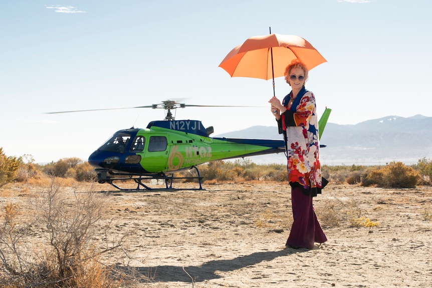 A woman in her late 60s with blonde hair and fancy outfit stands in a desert with an orange umbrella, a helicopter behind her