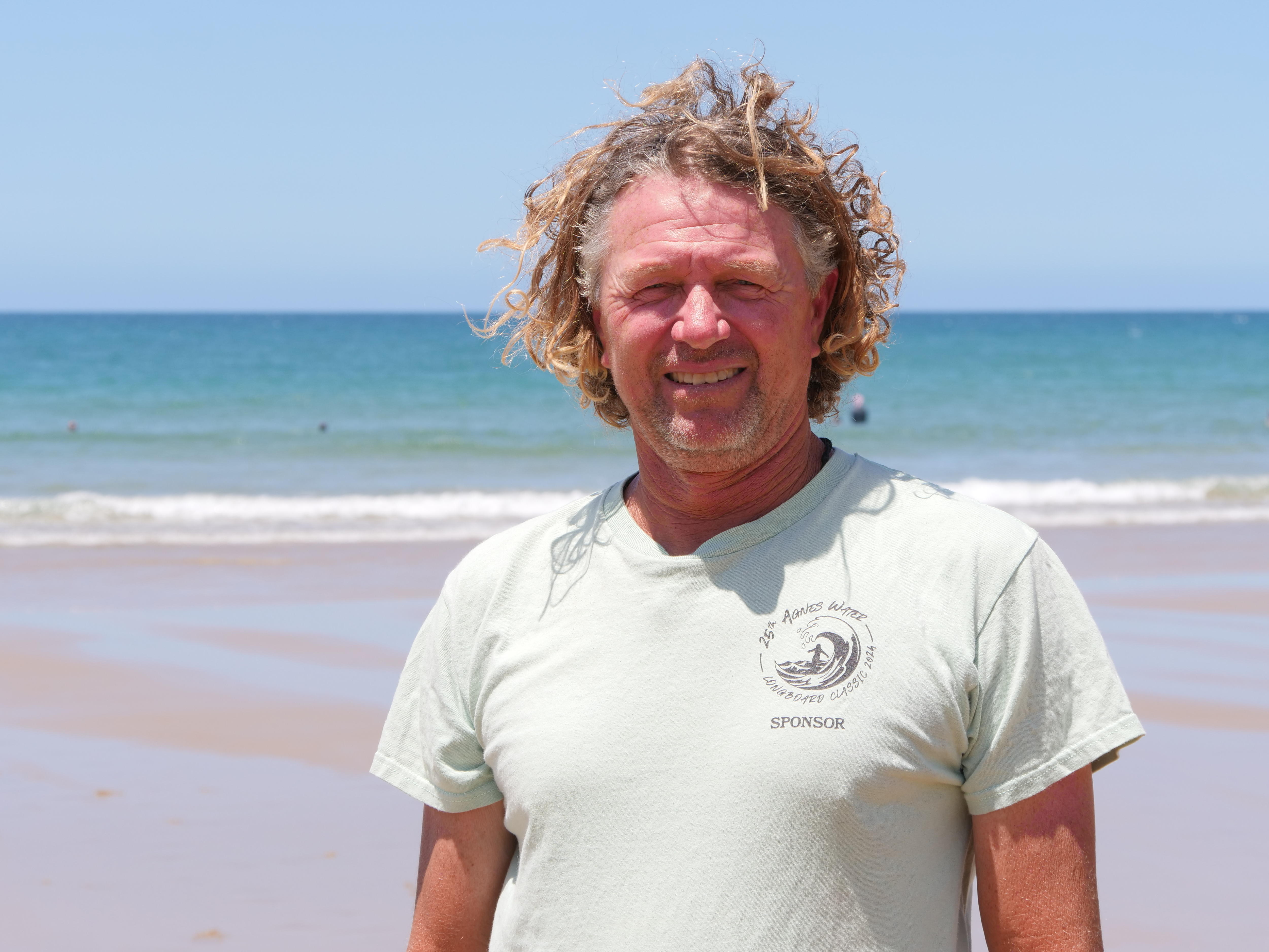 Paul White con rizos rubios, vestido con una camiseta y sonriendo directamente a la cámara, con la playa al fondo.