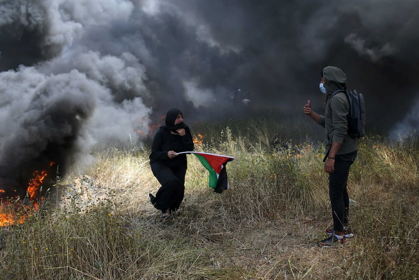 A woman is seen holding a Palestinian flag and surrounded by heavy smoke, while a young man gestures to her to move away.