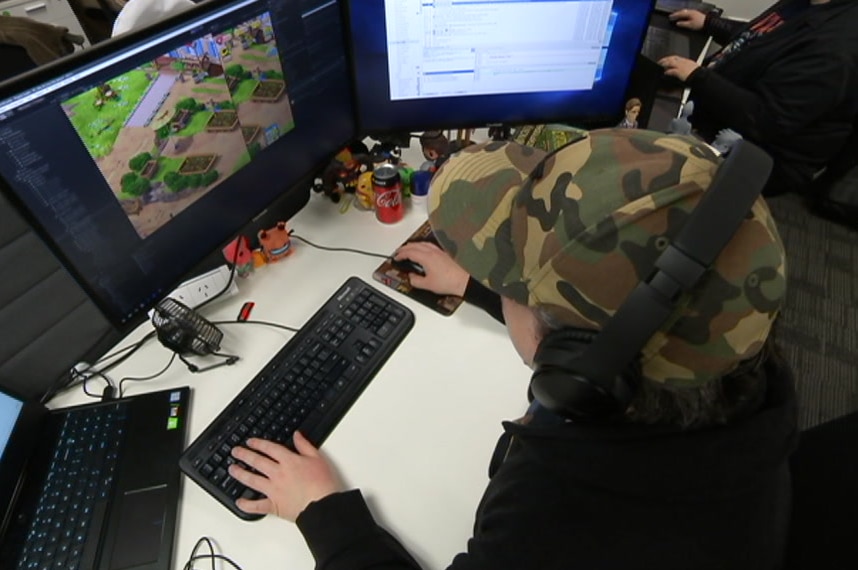 A person in a cap sits in front of a computer with a video game on the screen