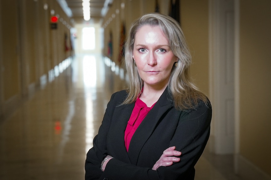 A young blonde woman in a black blazer stands with her arms crossed in a long hallway