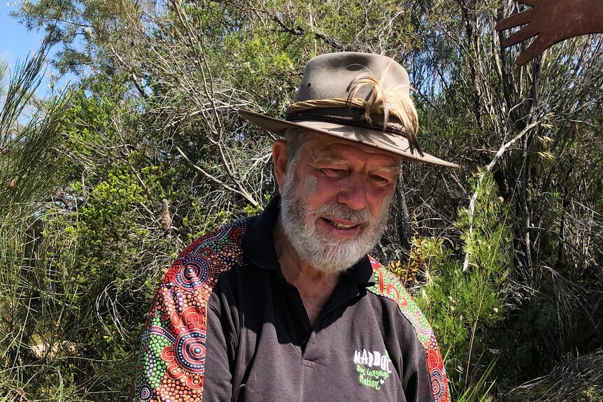 Middle aged man with beard wearing an akubra and NAIDOC shirt sitting on a rock in the bush