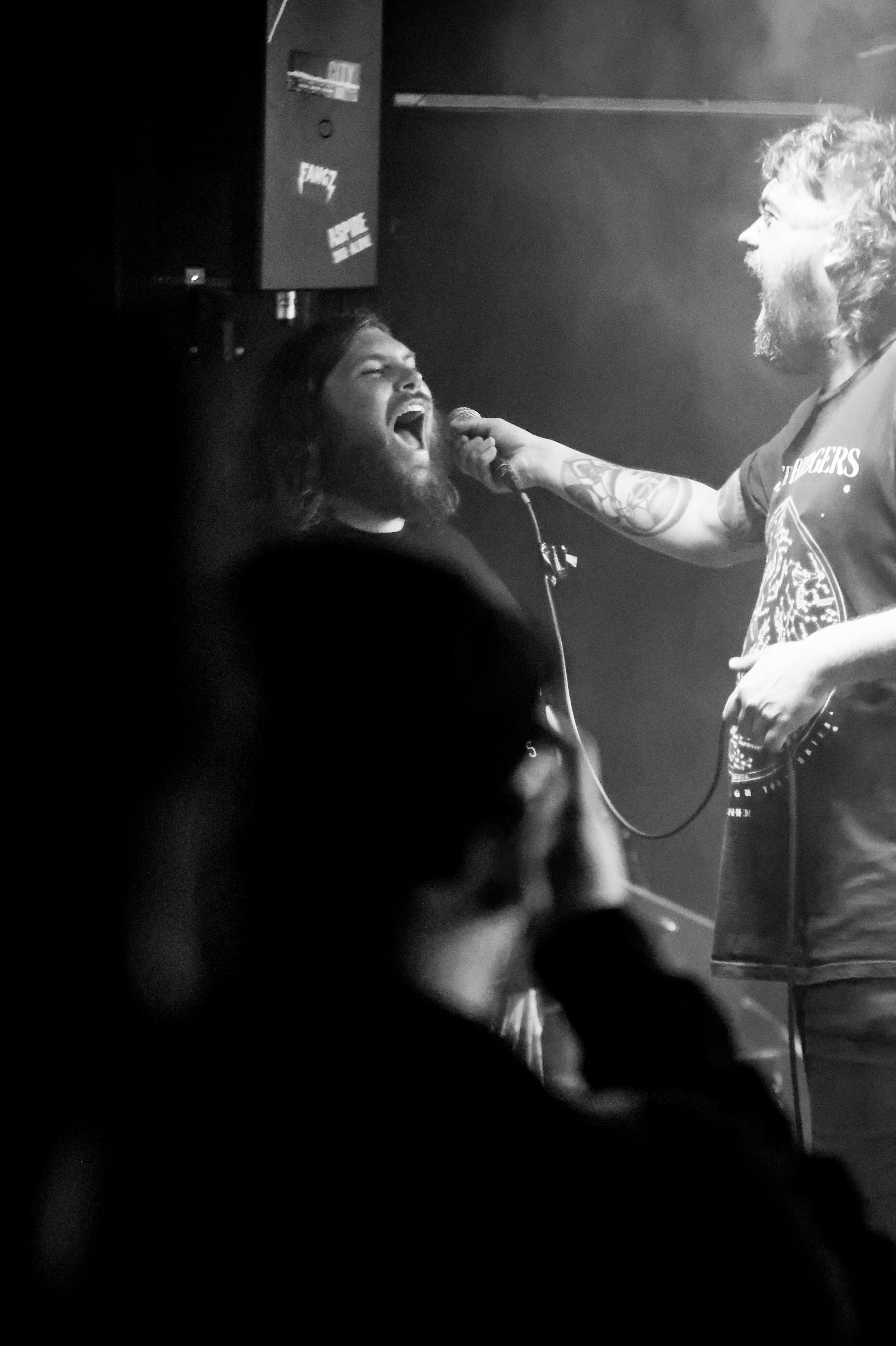A bearded man is onstage holding a microphone in front of another bearded man who is singing or yelling