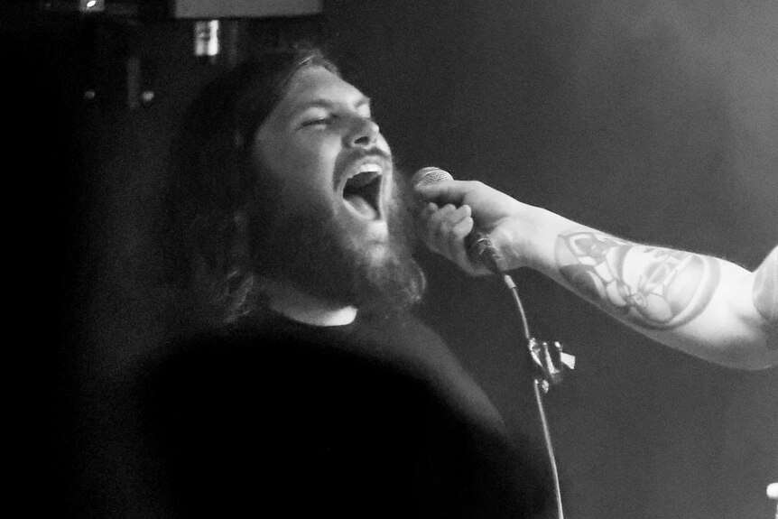 A bearded man is onstage holding a microphone in front of another bearded man who is singing or yelling