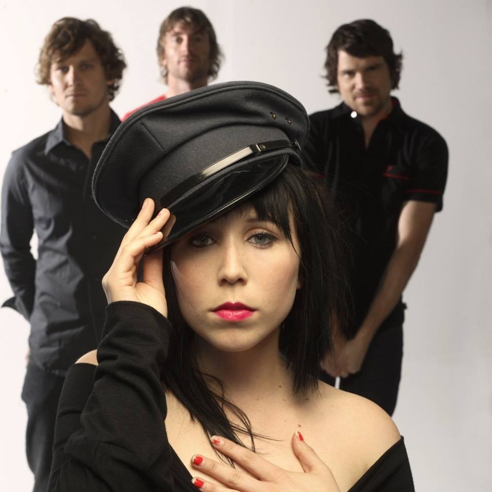 Foto de grupo com uma pessoa na frente usando chapéu escuro e outras em pé ao fundo.