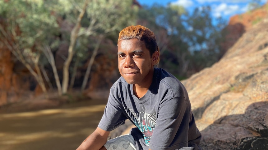 Leeroy is sitting on a rock by a river bank in alice springs wearing a dark shirt and white shorts smiling