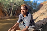 Leeroy is sitting on a rock by a river bank in alice springs wearing a dark shirt and white shorts smiling