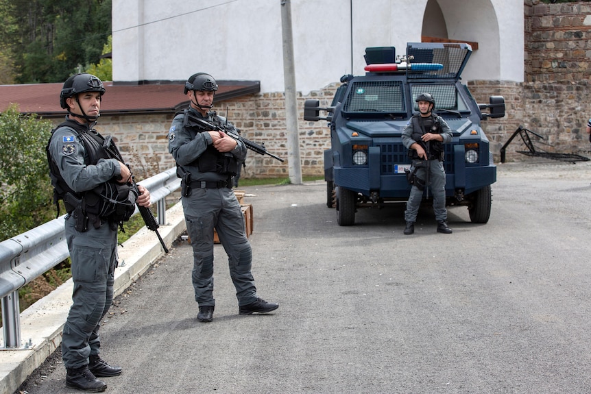 Three heavily armed police officers patrol a street with an armoured vehicle.