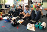 four students in blue uniforms sit on floor with books and pens in front of them engaging in lesson