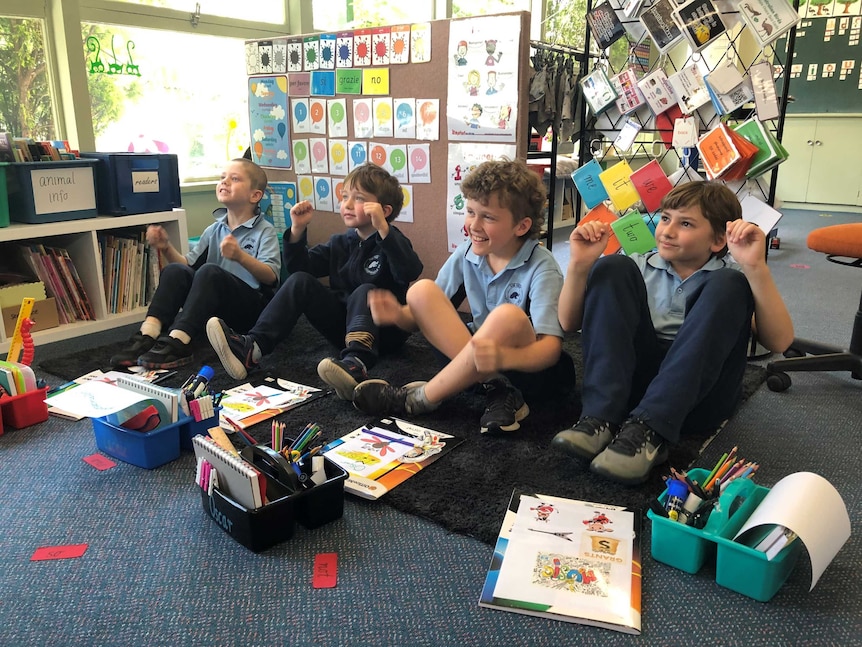 four students in blue uniforms sit on floor with books and pens in front of them engaging in lesson