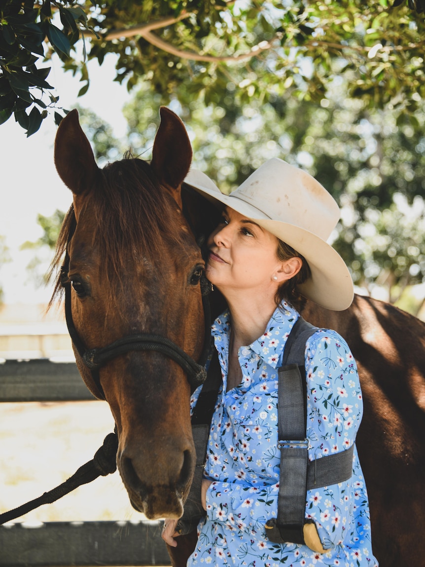 A woman with left arm in a sling and wearing a wide brim hat puts her face on a horse's face