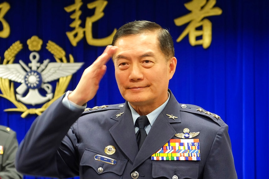 Shen Yi-ming raises his hand to his forehead as he salutes in military regalia in front of a bright blue background.