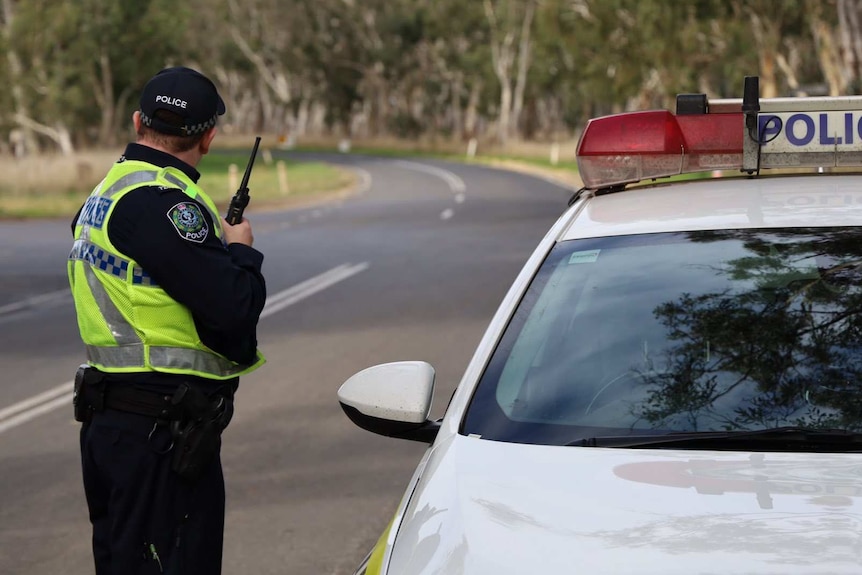 A police officer speaks into a radio next to a police car on a road with trees.