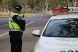 A police officer speaks into a radio next to a police car on a road with trees