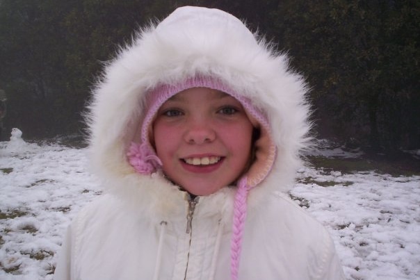 A young girl stands in the snow wearing a white fur hood.