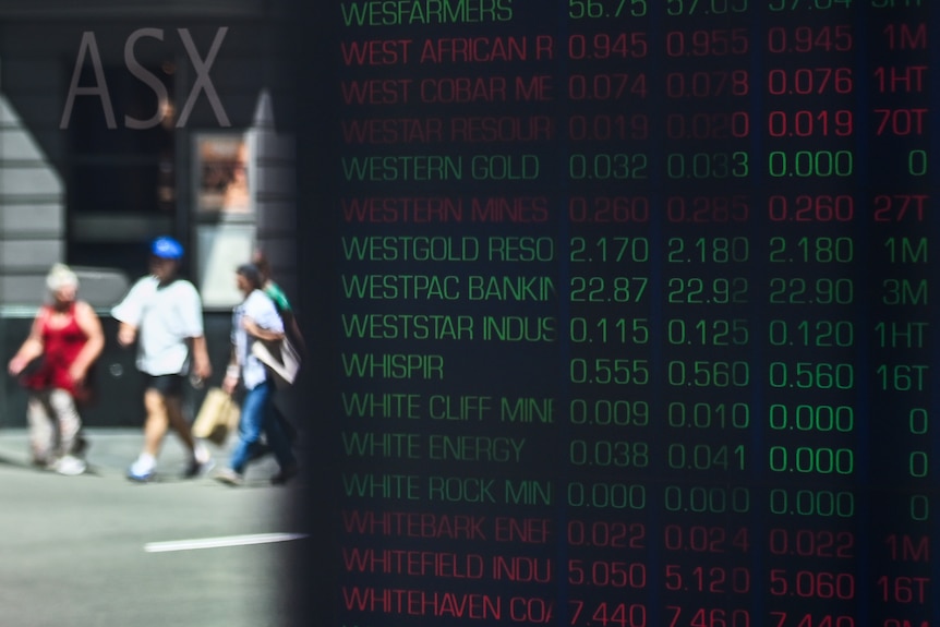 A close up of a board listening company names in green or red. Blurred pedestrians are in the background.