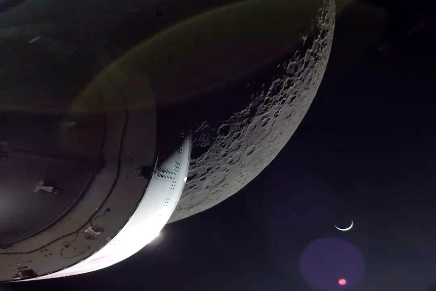 A spacecraft moves towards a cratered grey moon as it emerges from the darkness of space.