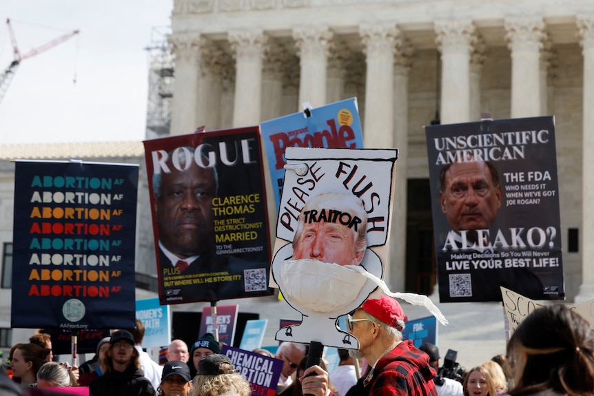 Signs are held up outside the Supreme Court with messages like 'Abortion AF' and 'Please Flush Traitor' with a photo of Trump 