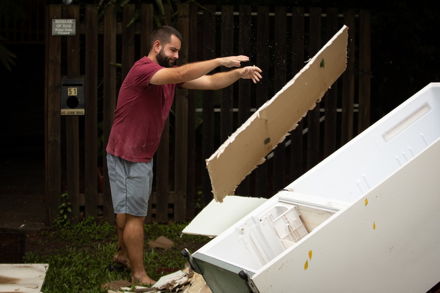 a man in a maroon shirt and shorts throws a piece of timber on to a pile of debris including a broken fridge