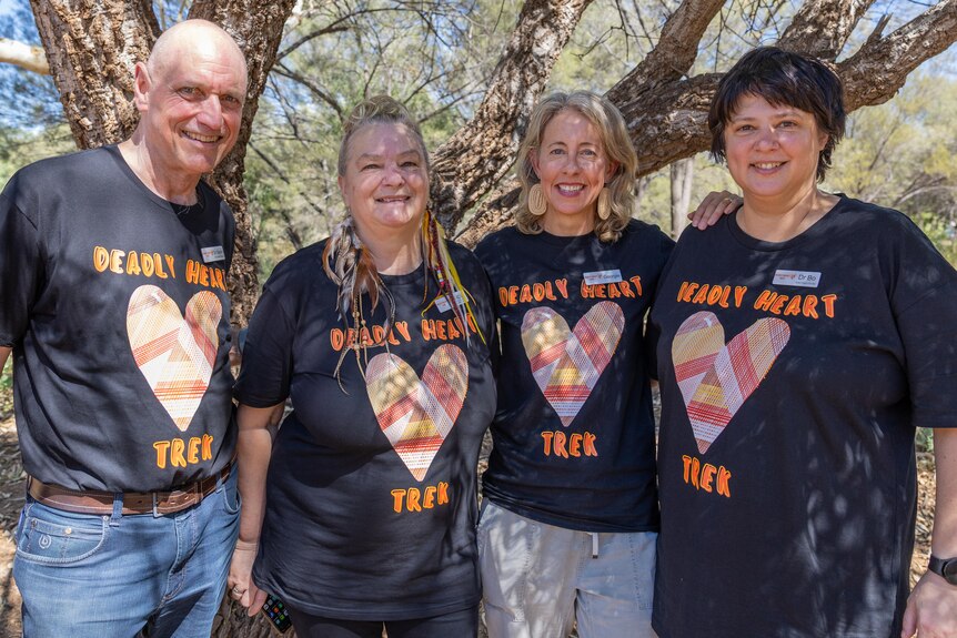 Four people stand in front of a tree. They are smiling, and wear black shirts which say Deadly Heart Trek on them.