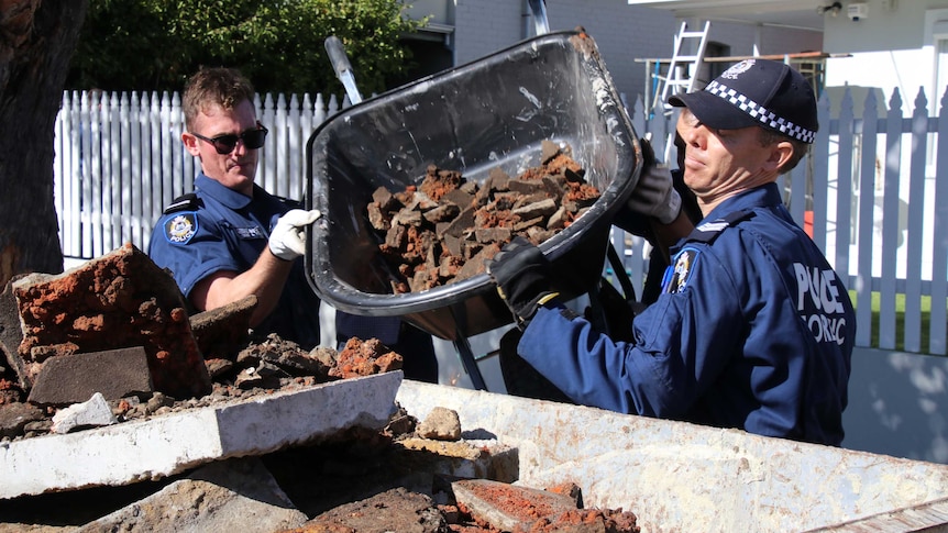 Two police forensic officers lift up a wheelbarrow carrying rubble to tip it into a skip bin outside a house.