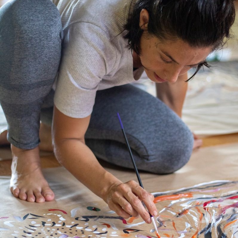 A dark-haired woman, barefoot, kneels over an artwork, dabbing t it with a paintbrush.