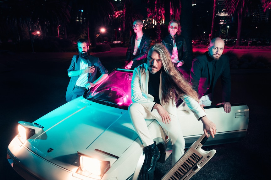 A band stand around or sit on a white sports car at night with its pop-up headlights on.