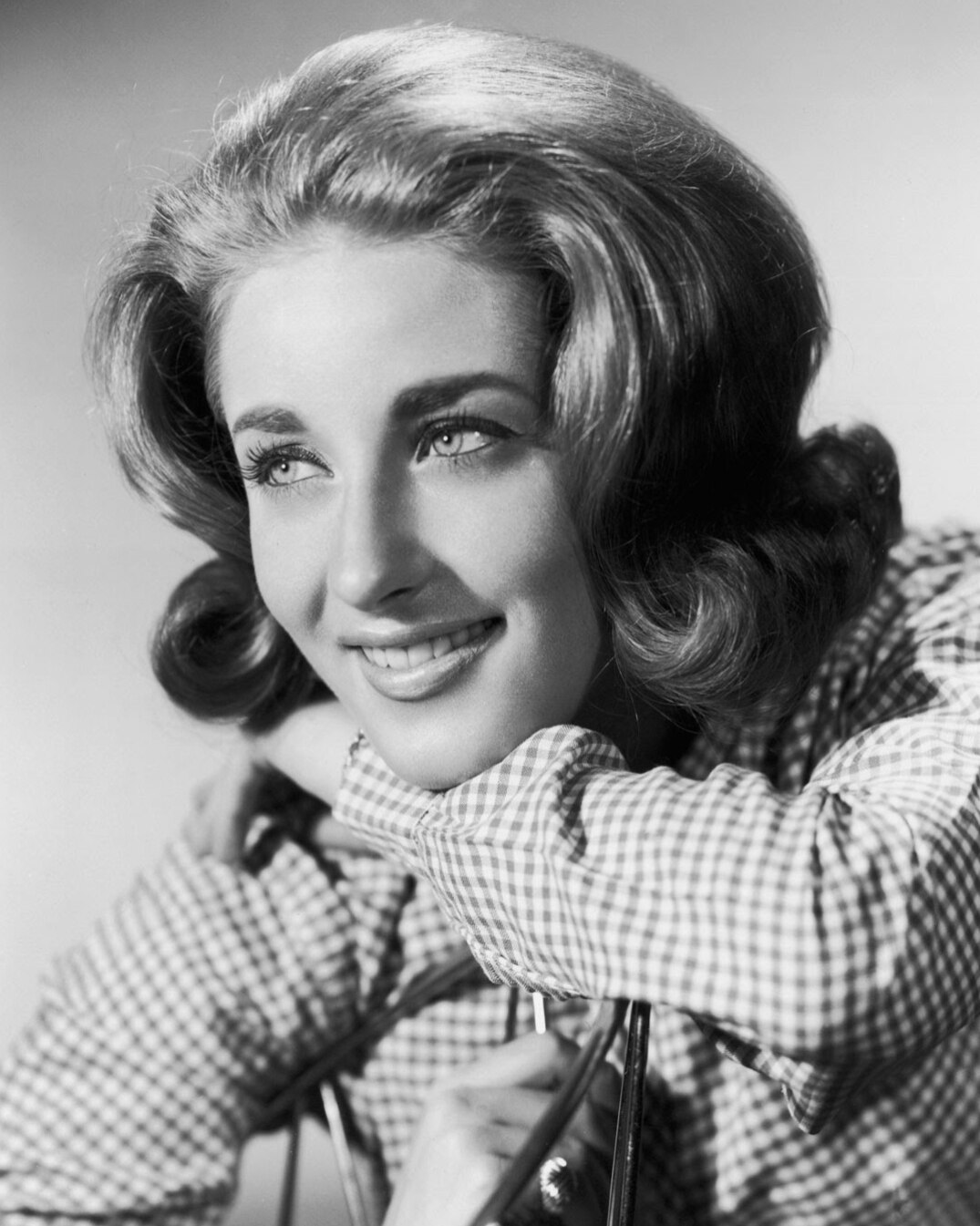 A young woman smiles. She has blonde, styled hair, wears a gingham shirt and rests her chin on her arm.