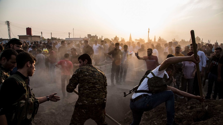 Clouds of dust fill the air as soldiers dig graves, surrounded by a huge crowd next to a road.